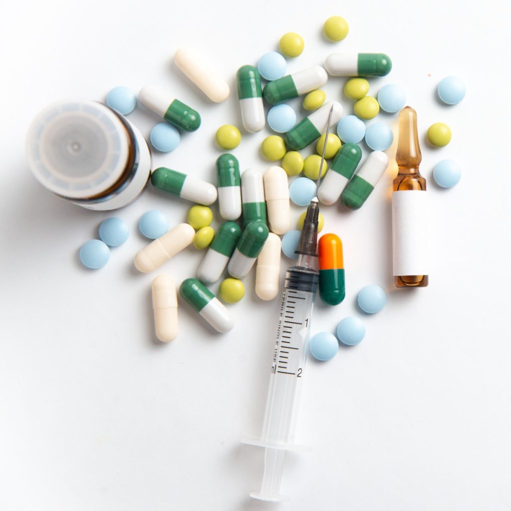 Bottle, ampoule, syringe and several multi-coloured pills on white backdrop.